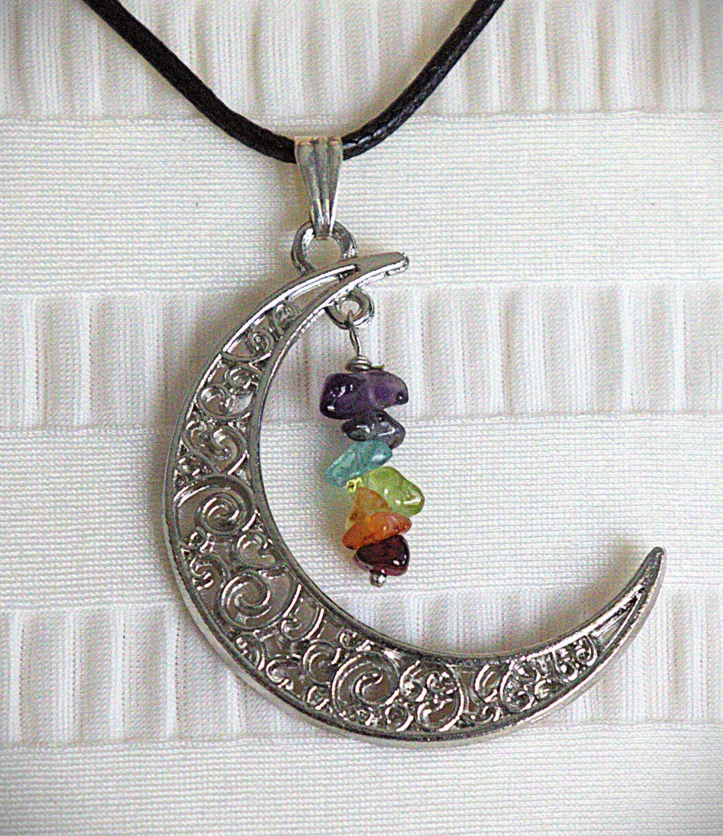 A silver crescent moon pendant necklace with intricate scrollwork, hanging from a black cord. The pendant is decorated with a small cluster of colorful gemstone beads in purple, blue, green, orange, and red, set against a white textured fabric background.