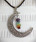 A silver crescent moon pendant necklace with intricate scrollwork, hanging from a black cord. The pendant is decorated with a small cluster of colorful gemstone beads in purple, blue, green, orange, and red, set against a white textured fabric background.