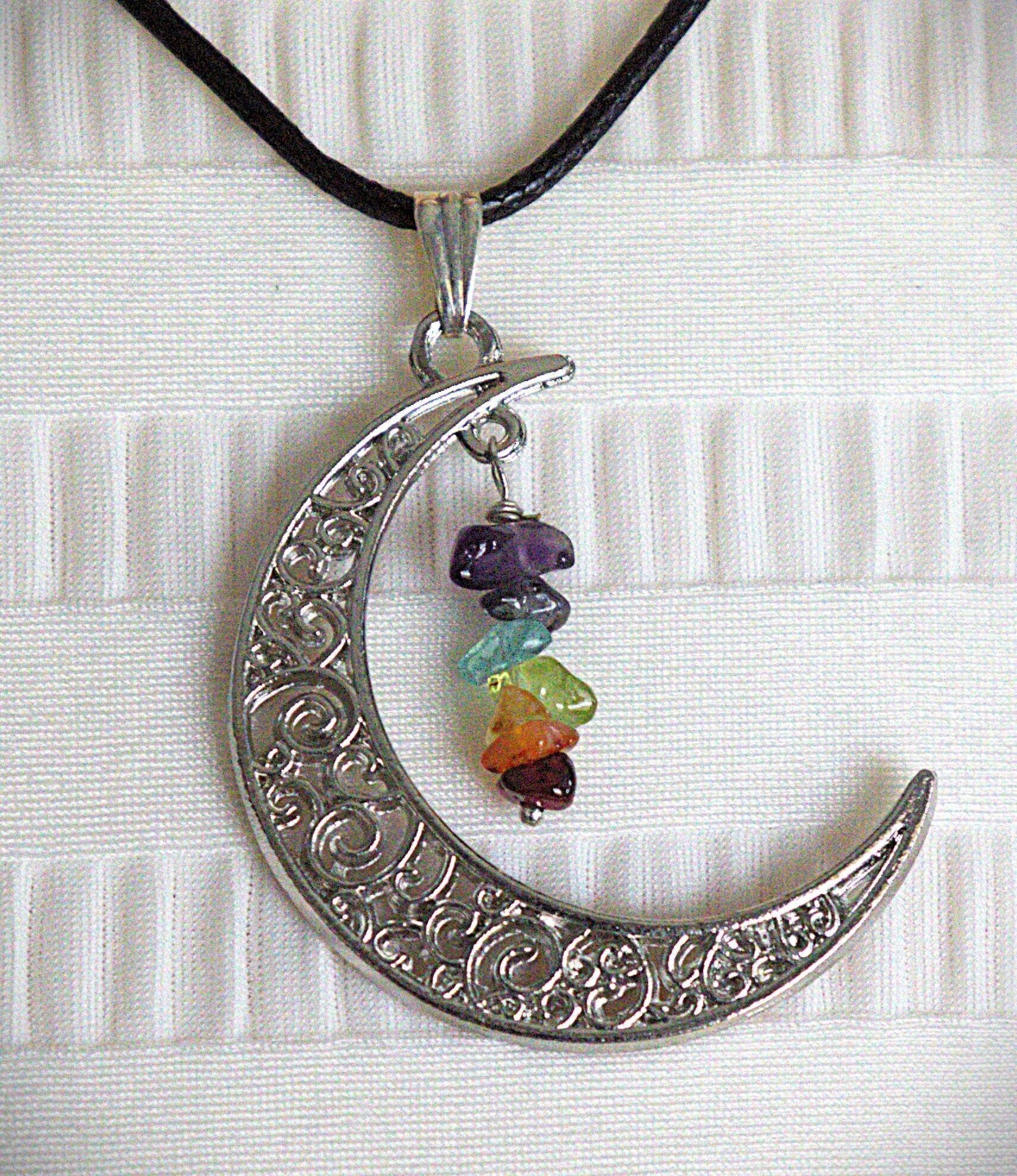 A silver crescent moon pendant necklace with intricate scrollwork, hanging from a black cord. The pendant is decorated with a small cluster of colorful gemstone beads in purple, blue, green, orange, and red, set against a white textured fabric background.