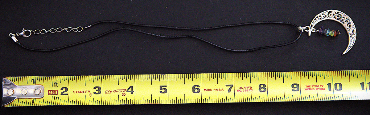 A black cord necklace with a silver crescent moon pendant and a small multicolor bead charm, shown above a yellow tape measure on a black background.