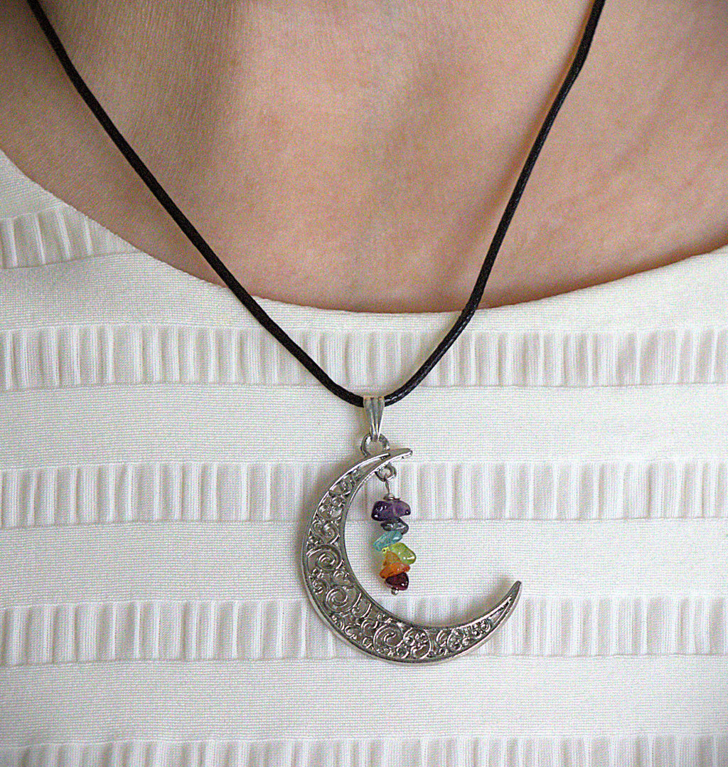 A close-up of a person wearing a silver crescent moon pendant necklace with a black cord and small multicolored gemstone beads, over a white ribbed top.