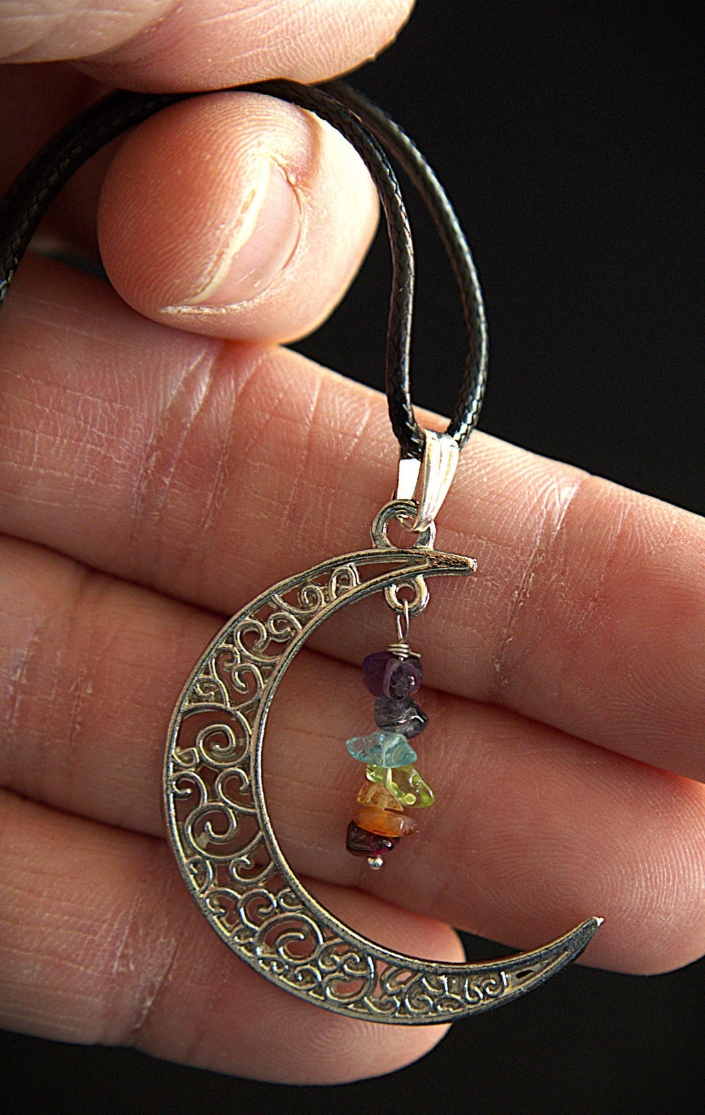 A close-up of a hand holding a crescent moon pendant necklace. The pendant is made of silver-toned metal with intricate filigree cutouts and hangs from a black cord.