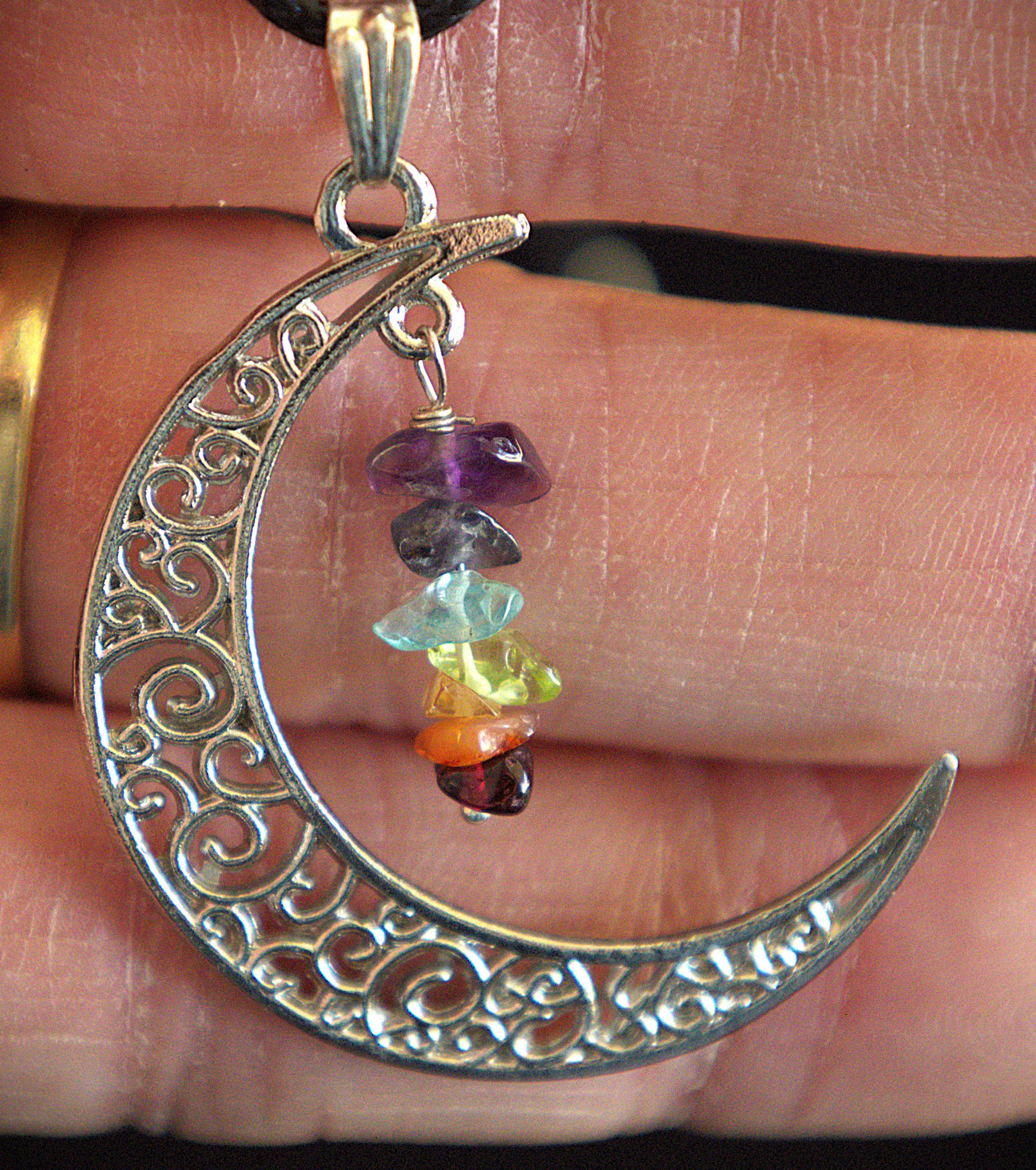 A silver crescent moon pendant with intricate scrollwork, featuring a dangling charm made of stacked multicolored gemstone chips, held in a person's hand.