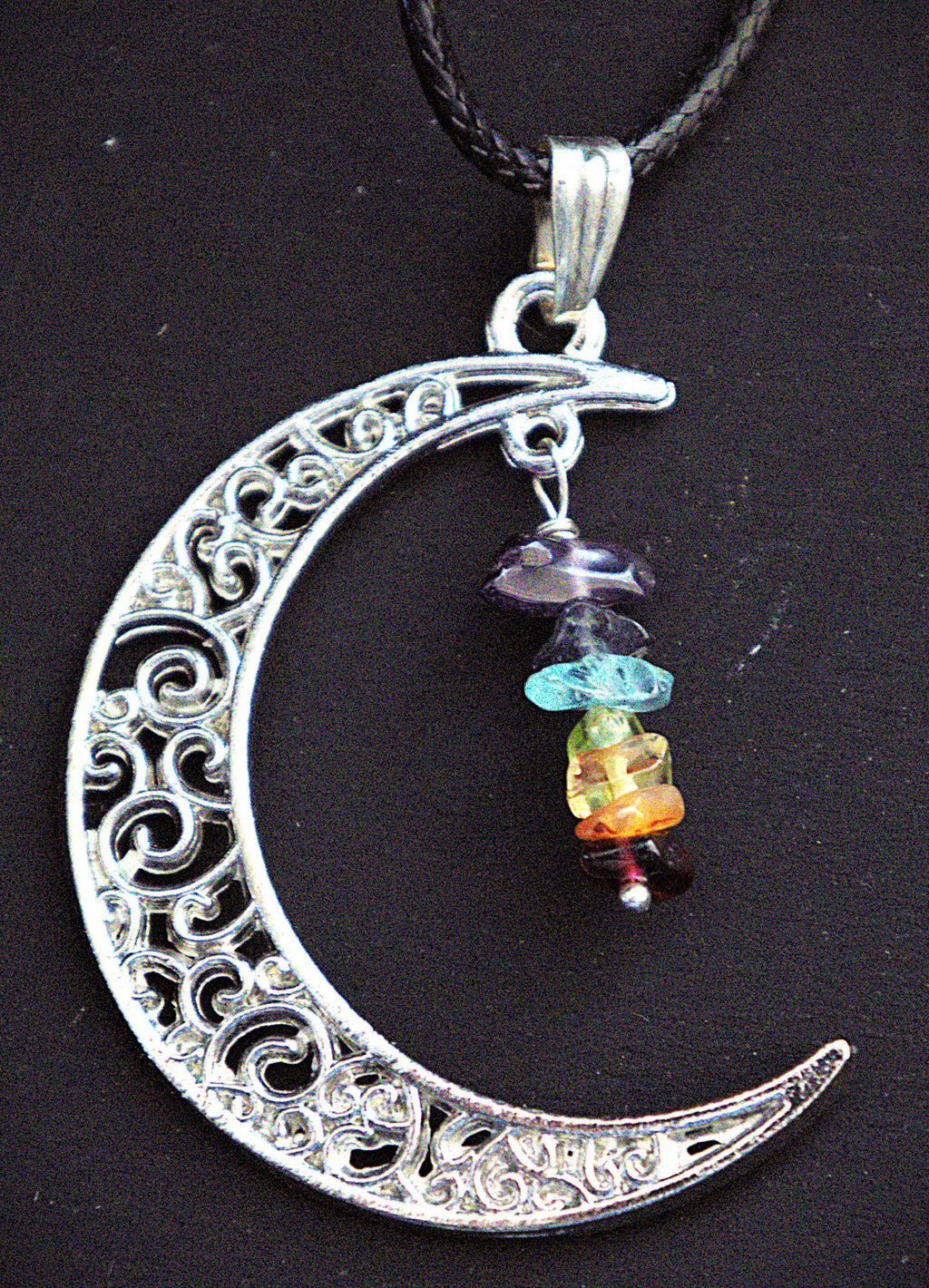 A silver crescent moon pendant necklace with intricate filigree cutouts and a dangling multi-colored gemstone bead charm, shown on a black cord against a dark background.