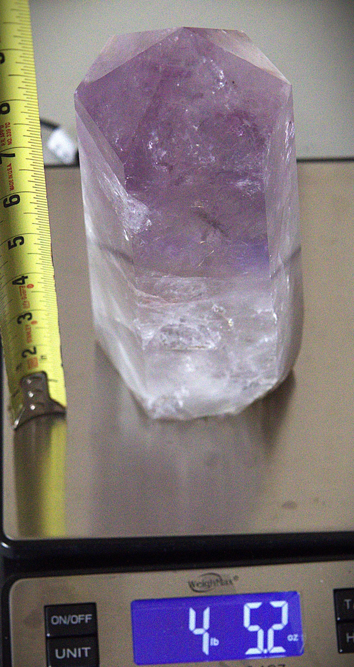 A raw amethyst crystal cluster with a purple top section and a clear white bottom, resting on a digital scale next to a yellow measuring tape.