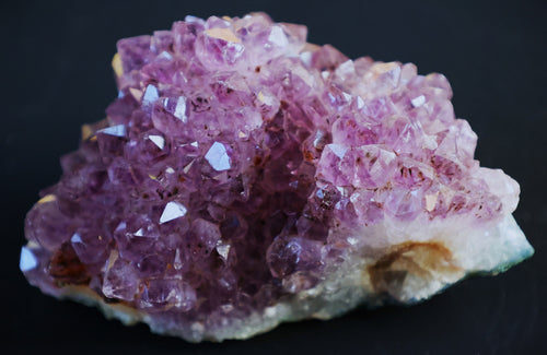 A natural amethyst crystal cluster with prominent purple and pink crystalline formations on a light-colored matrix, photographed against a solid black background.