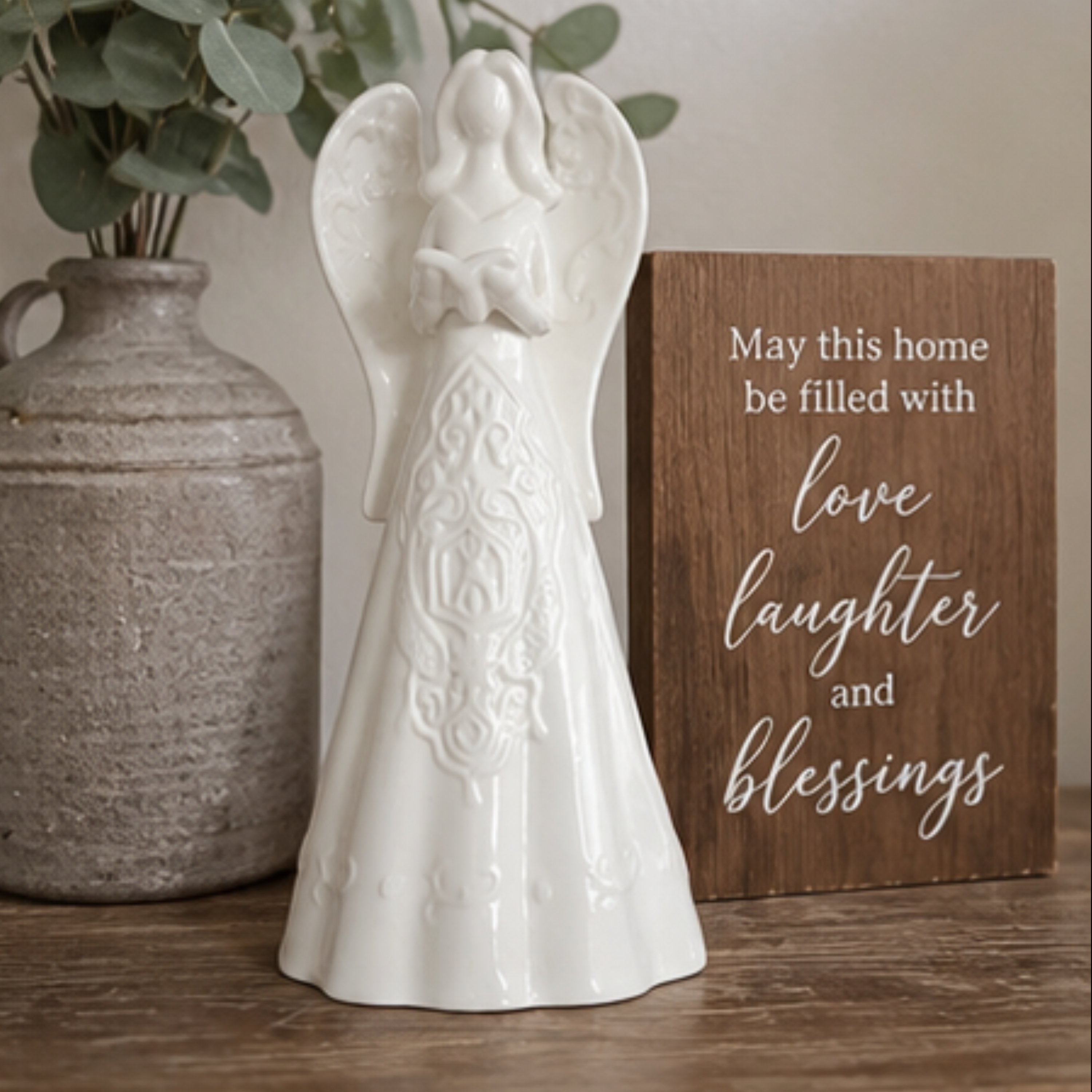 A white ceramic angel figurine with a child on its shoulders stands on a wooden surface beside a brown wooden sign that reads, 'May this home be filled with love, laughter and blessings.' A textured gray vase with greenery is visible in the background.