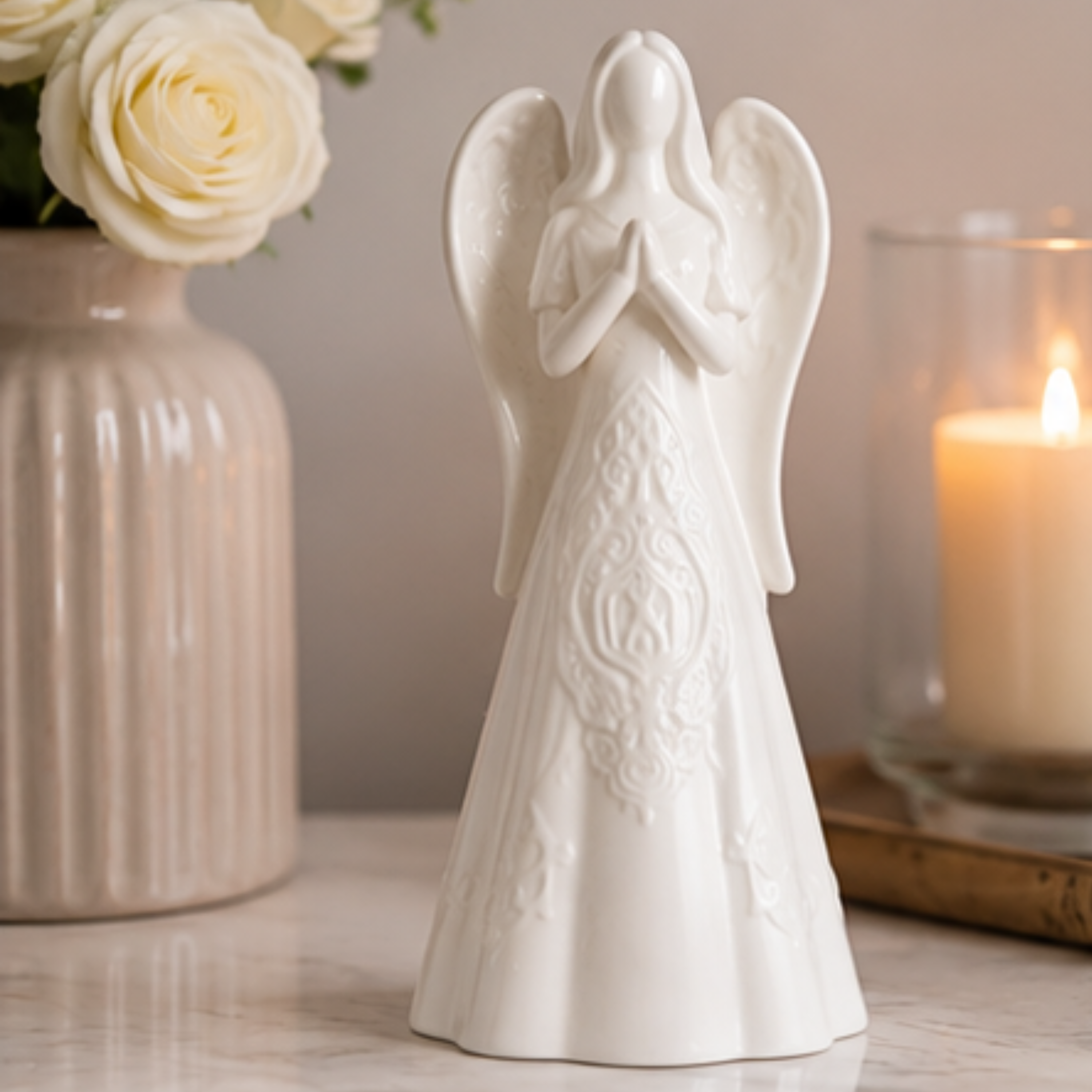 A white ceramic angel figurine with wings and a prayer pose stands on a tabletop, styled with a vase of white roses on the left and a lit candle in a glass holder on the right.