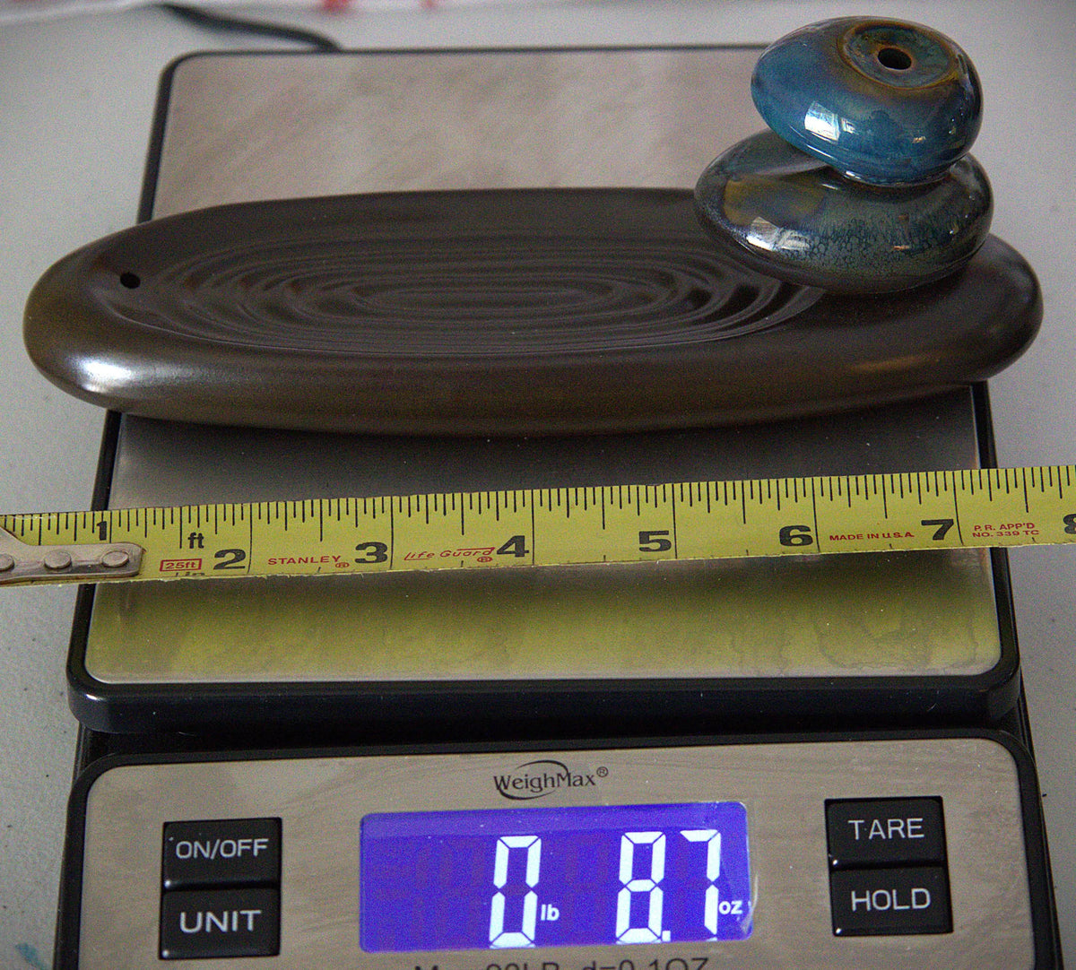A dark brown carved wooden tray or board with concentric grooved patterns, topped with two stacked blue-green polished stones, shown on a digital scale with a yellow tape measure in front.