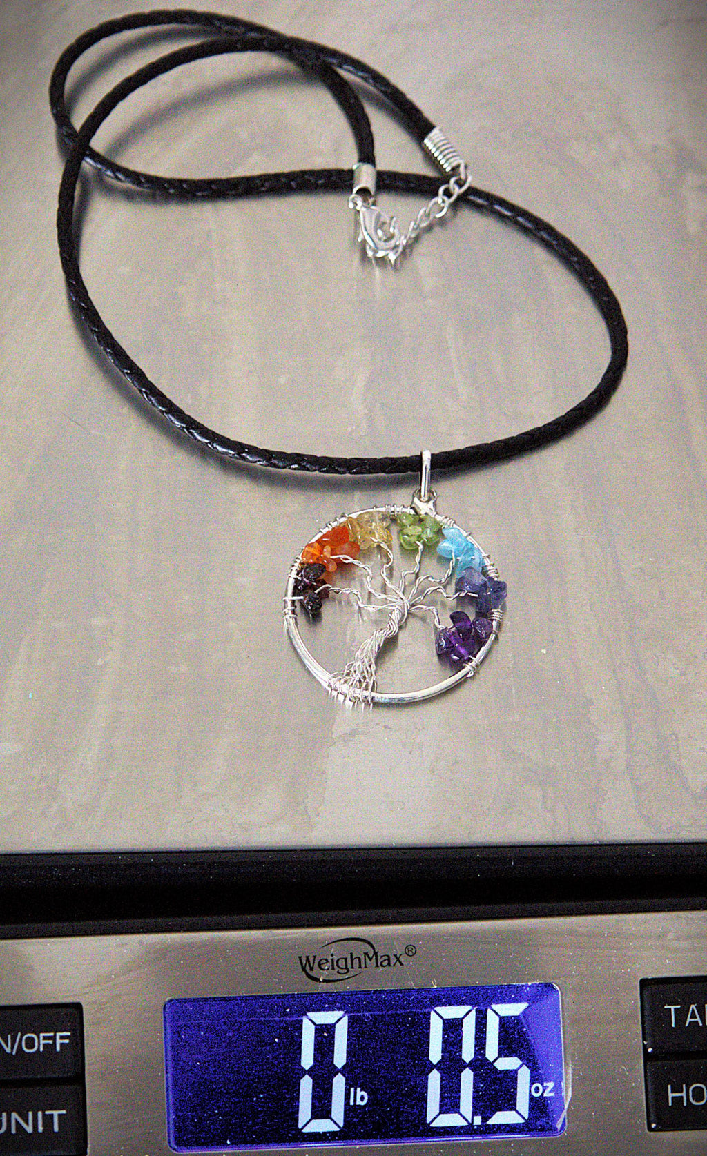 A black cord necklace with a silver tree of life pendant set with rainbow-colored gemstones, shown resting on a digital scale displaying 0.05 oz.