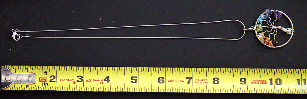 A silver-toned tree of life pendant necklace with a multicolor circular pendant, shown on a black background with a yellow measuring tape below for scale.