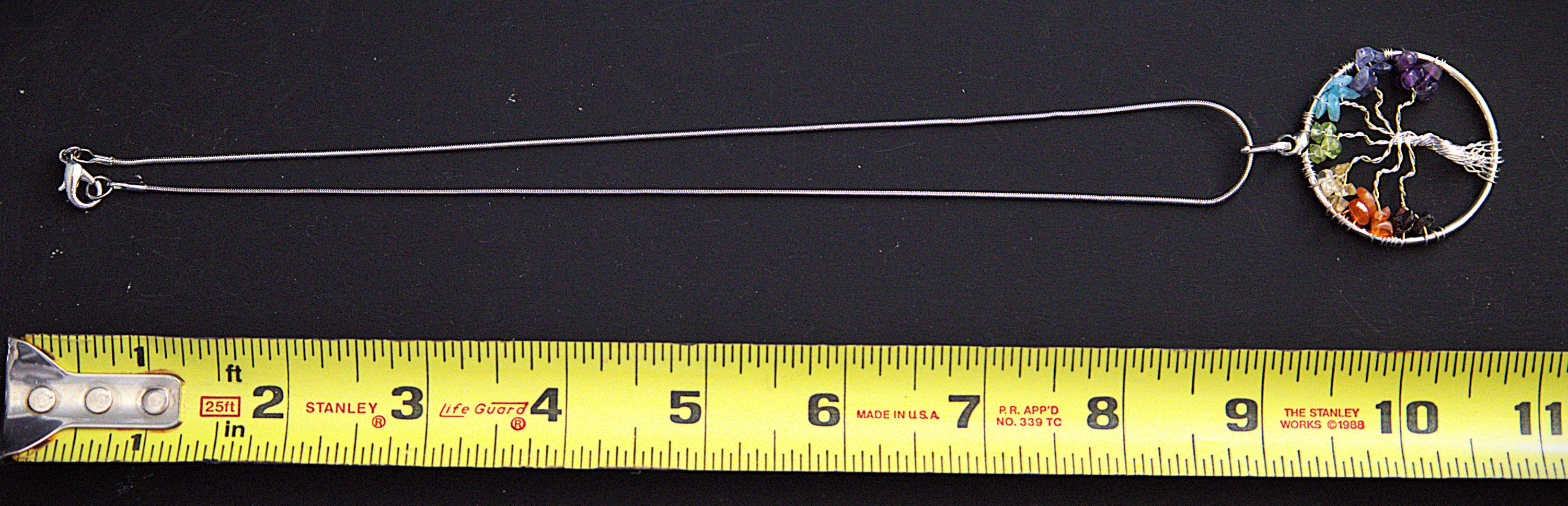 A silver-toned tree of life pendant necklace with a multicolor circular pendant, shown on a black background with a yellow measuring tape below for scale.