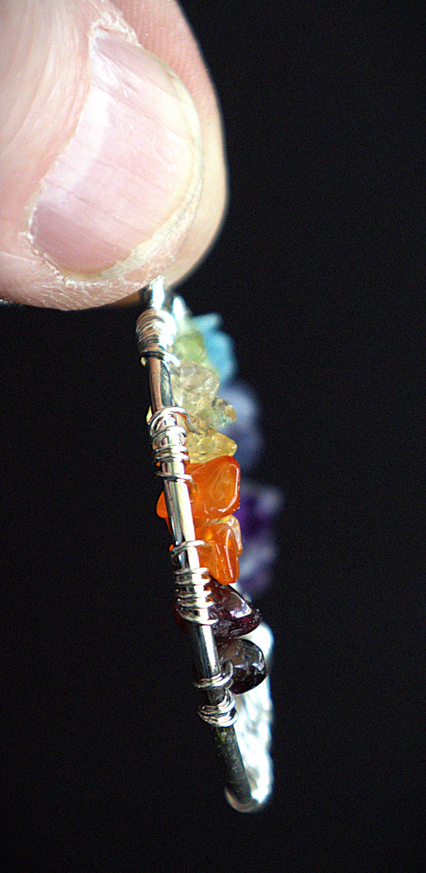 A close-up of a hand holding a silver-toned beaded charm or earring with multiple colored stones, including orange, purple, green, and blue, against a solid black background.