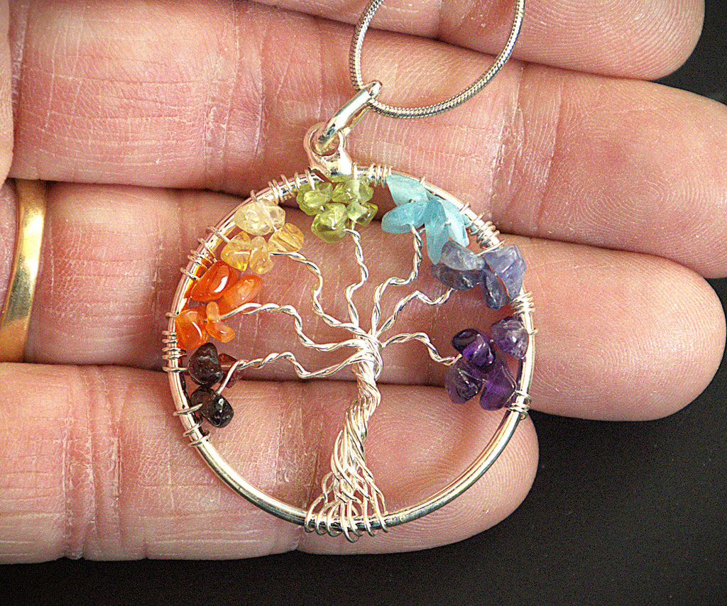 A silver tree of life pendant with colorful gemstone accents, held in a person's hand against a dark background.