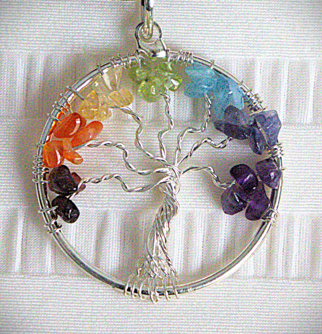 A circular silver wire tree of life pendant with seven colorful gemstone chakra clusters along the top and side borders, displayed on a white textured fabric background.