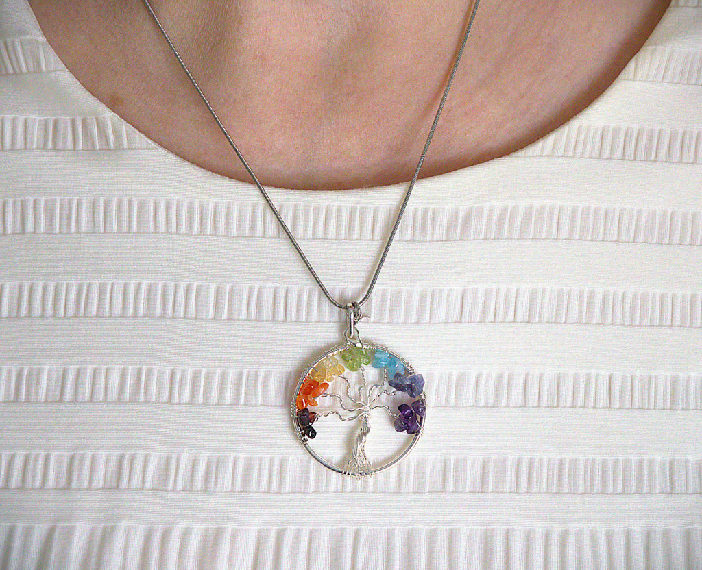 A silver tree of life pendant necklace with seven colorful gemstones, worn on a silver chain over a white textured top.