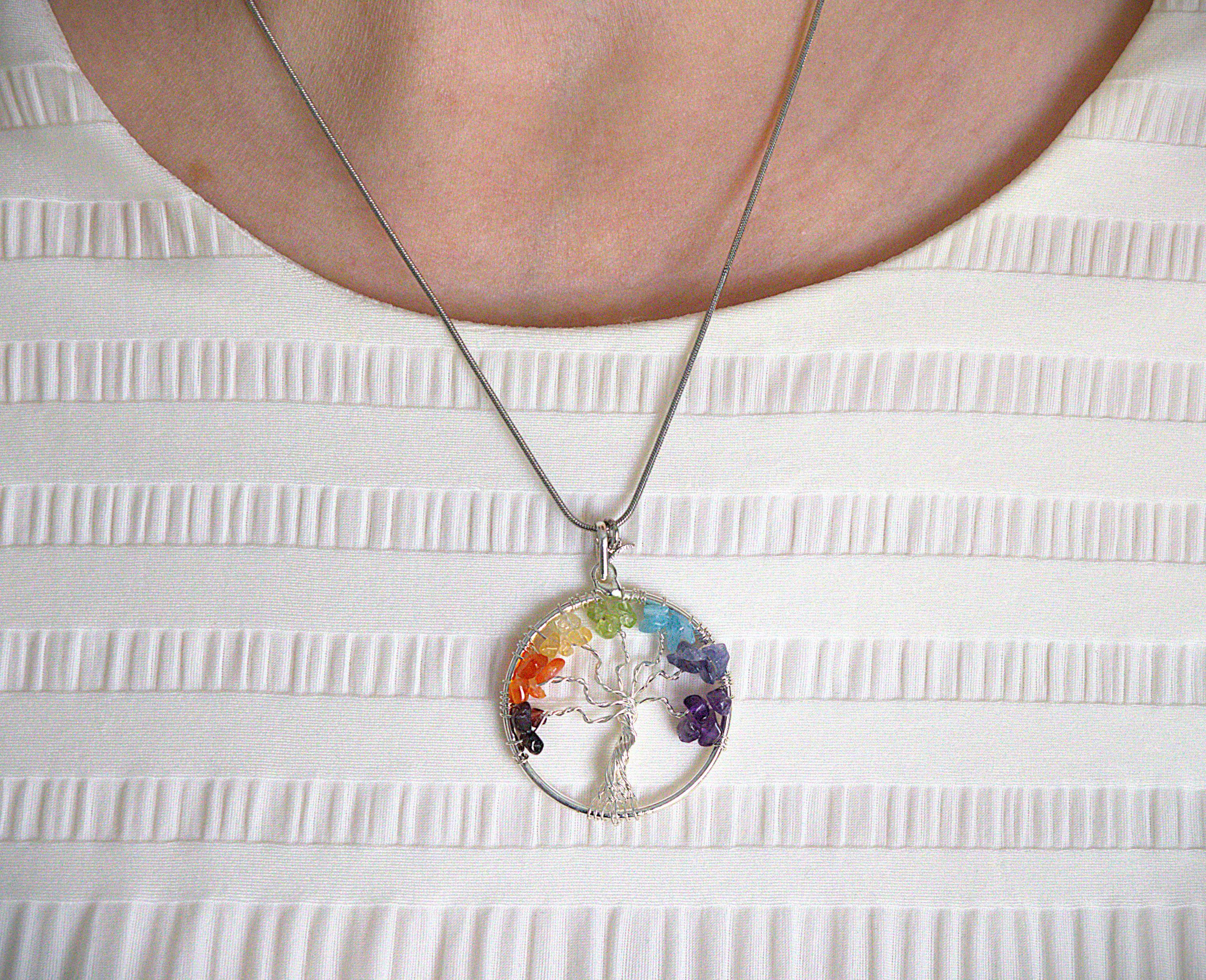 A silver tree of life pendant necklace with seven colorful gemstones, worn on a silver chain over a white textured top.