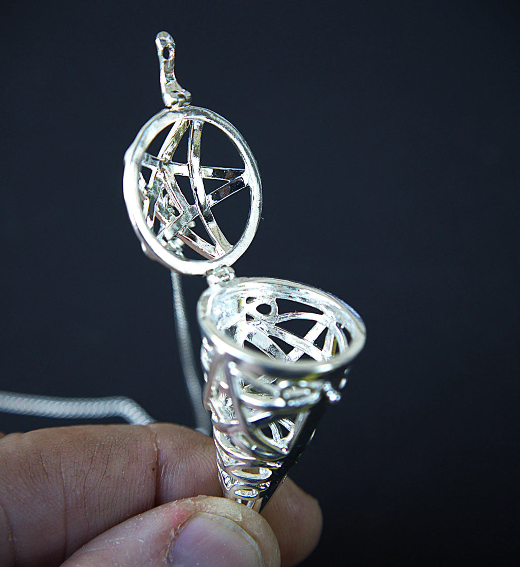 A silver-toned metal pendant held open by a person's fingers against a dark black background. The pendant features an open top section with an intricate pentagram design inside a circular frame, connected to a conical lower section with openwork geometric detailing and a chain.