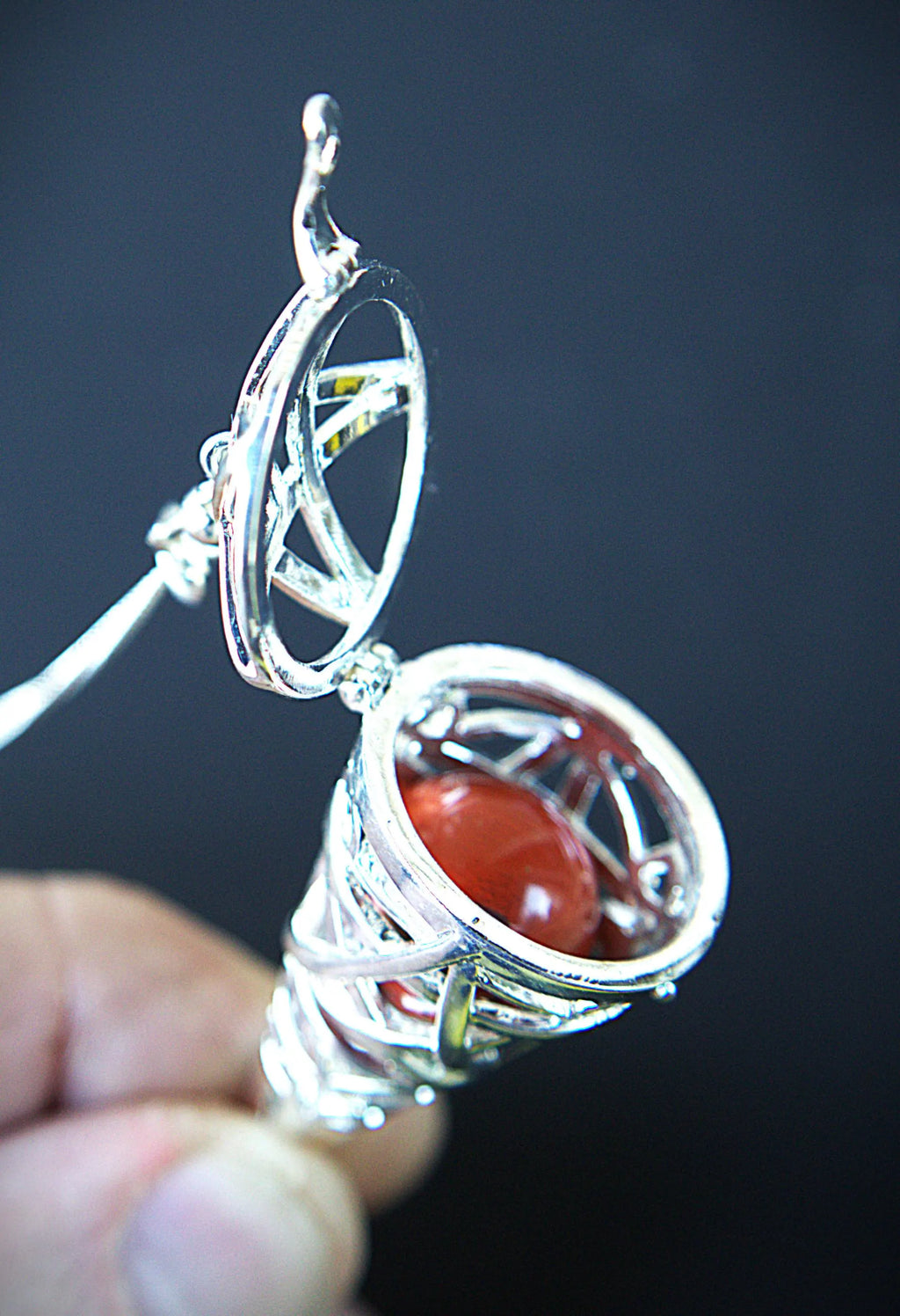 A close-up of a hand holding a silver-toned ring with a circular openwork top and a red cabochon gemstone in a textured basket-like setting, against a dark background.