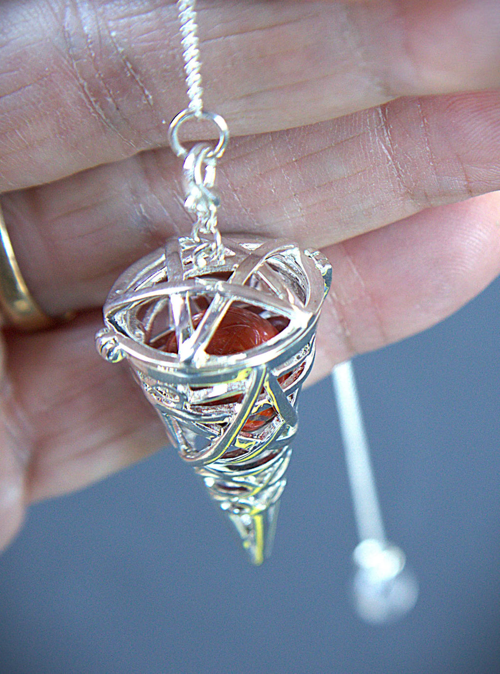 A silver-toned metal pentacle pendant held in a hand, featuring a red-orange stone set inside a conical cage with a silver pentagram design, suspended from a silver chain.