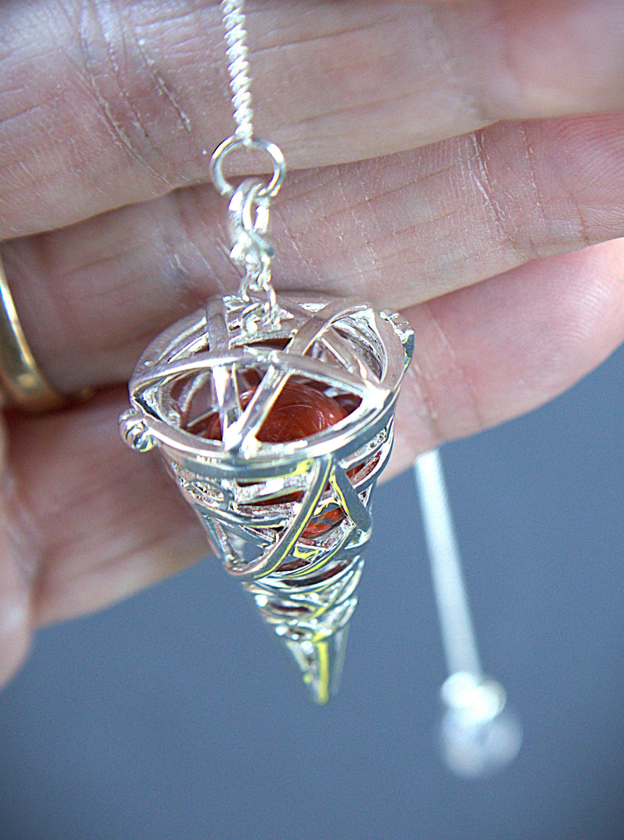 A silver-toned metal pentacle pendant held in a hand, featuring a red-orange stone set inside a conical cage with a silver pentagram design, suspended from a silver chain.