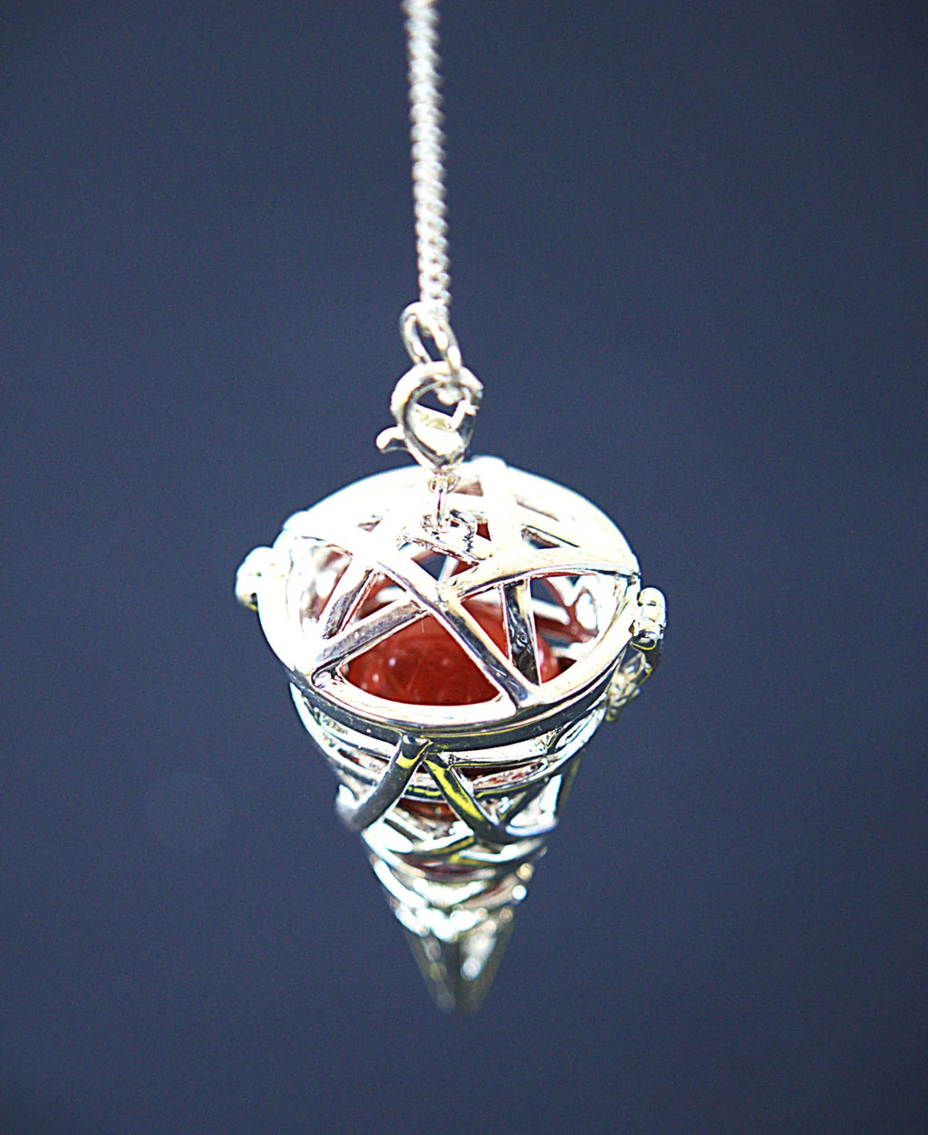 A silver-toned pendant necklace featuring a spherical cage-like structure enclosing a translucent red-orange stone, suspended from a fine chain against a dark blue background.