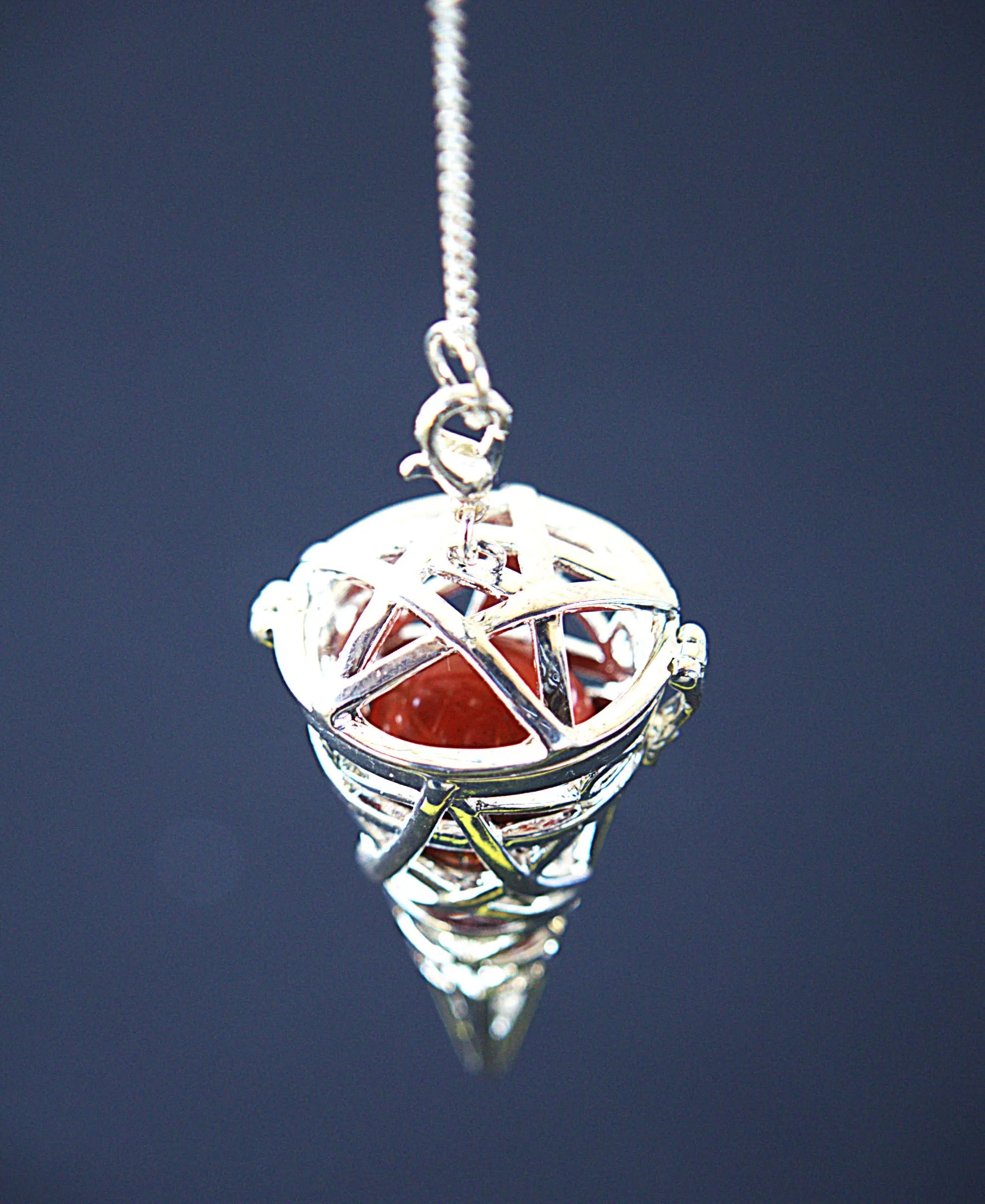 A silver-toned pendant necklace featuring a spherical cage-like structure enclosing a translucent red-orange stone, suspended from a fine chain against a dark blue background.