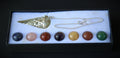 A crystal chakra pendant necklace set in a black gift box with a white insert. The set includes a gold-toned metal cone pendant and a silver-toned chain, with seven round cabochon gemstones in blue, pink, red, purple, yellow, brown, and green aligned along the bottom.