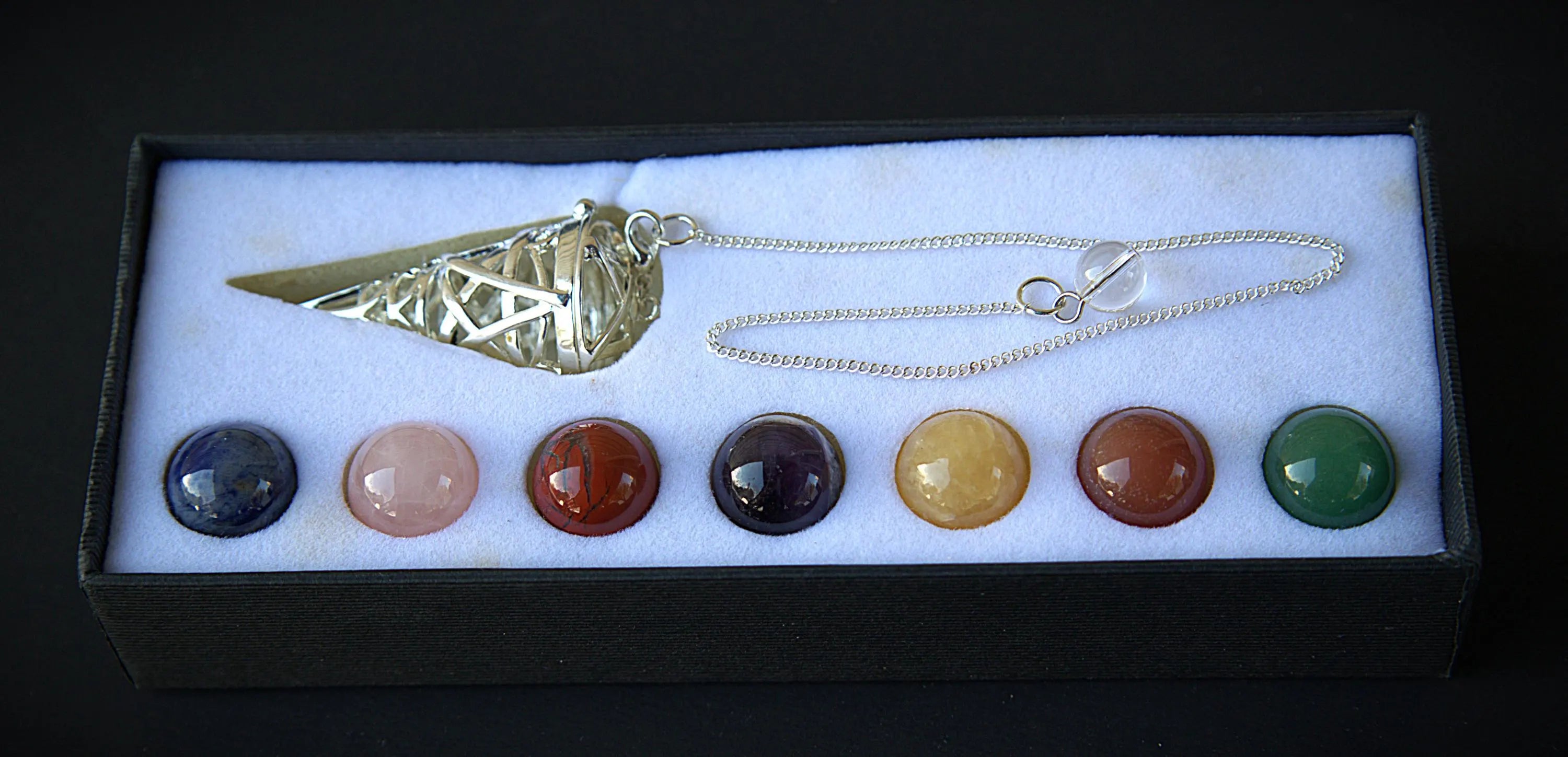 A crystal chakra pendant necklace set in a black gift box with a white insert. The set includes a gold-toned metal cone pendant and a silver-toned chain, with seven round cabochon gemstones in blue, pink, red, purple, yellow, brown, and green aligned along the bottom.