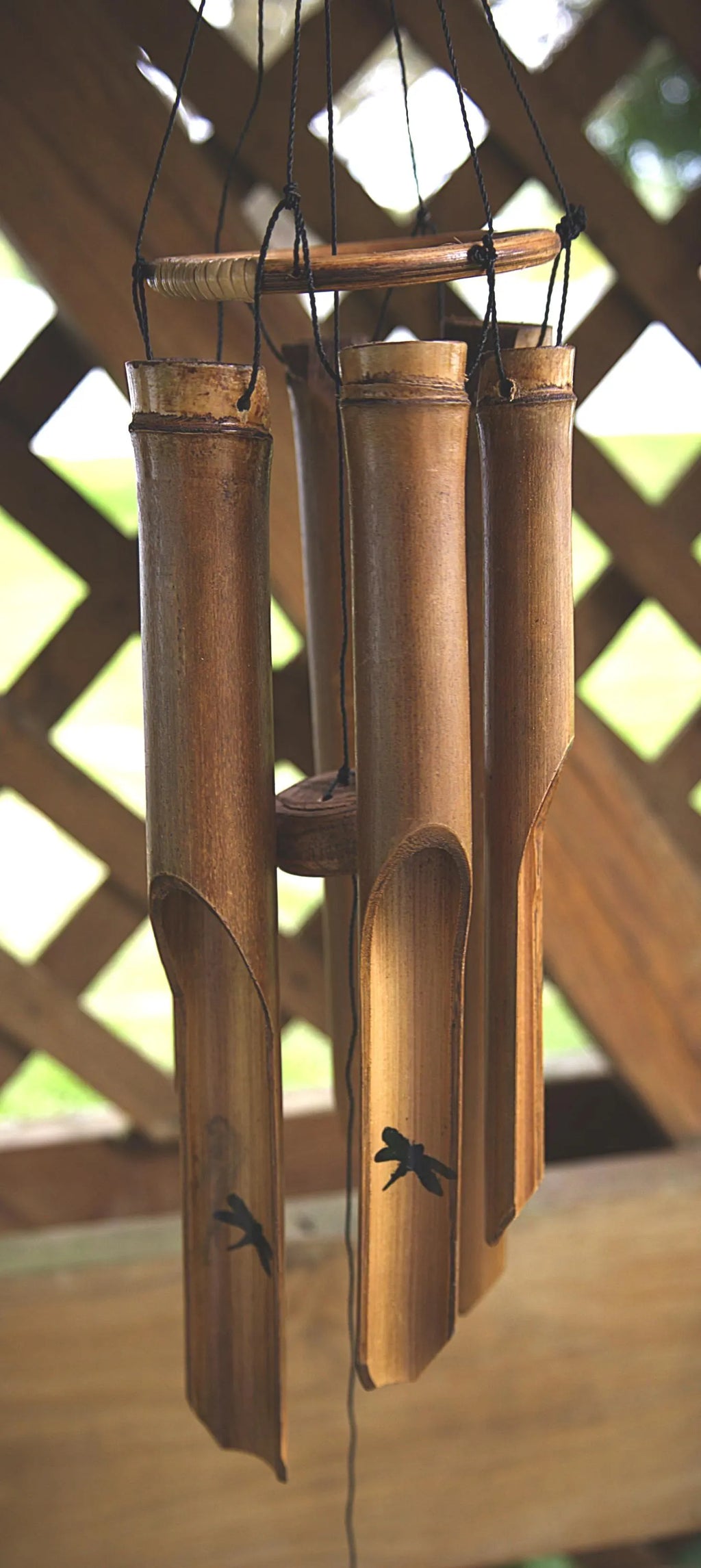 Three natural bamboo wind chimes hang from a wooden frame on a porch, with a blurred lattice background and greenery visible behind them.