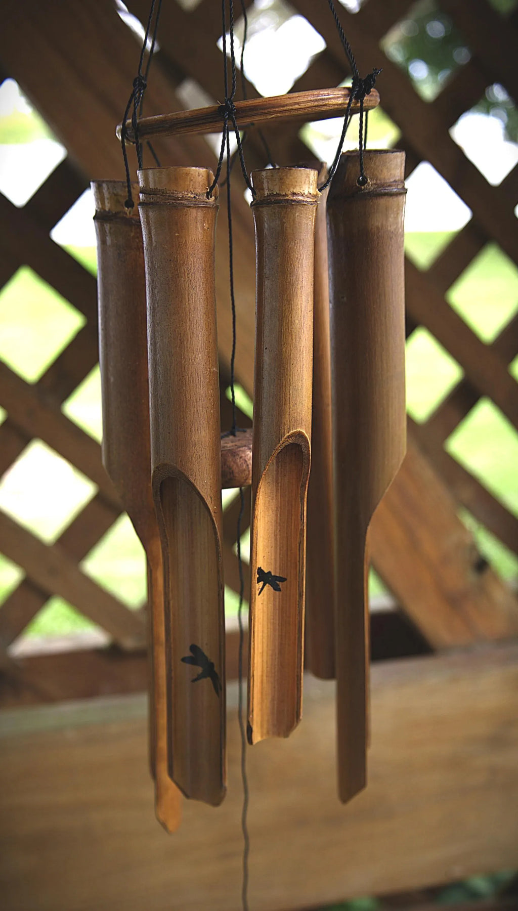 Bamboo wind chimes hanging from a wooden support structure, with a blurred outdoor lattice background.