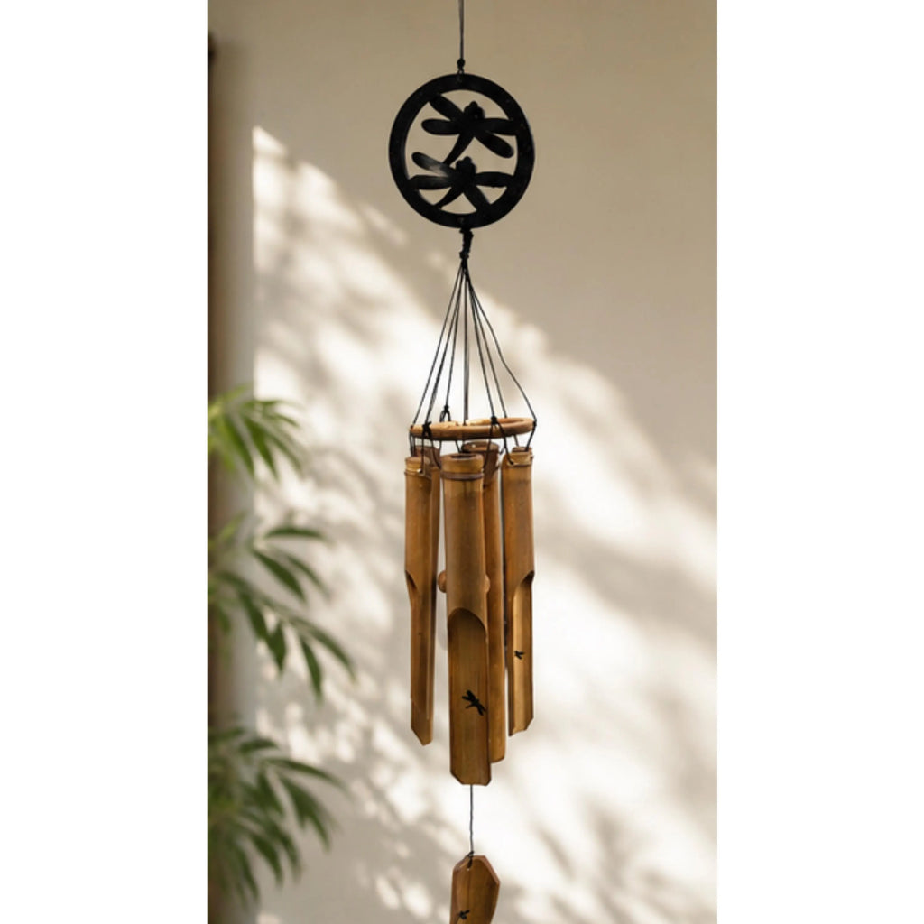 A bamboo wind chime hanging from a black circular metal top, set against a warm beige wall with soft leaf shadows and a hint of green foliage on the left.