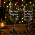 Two clear wine glasses with black stems and white engraved text reading 'Dear Santa, Bring Wine' and 'Dear Santa, Live Been Good - ish' with snowflake details, displayed on a wooden table with a Christmas tree and holiday decorations in the background.
