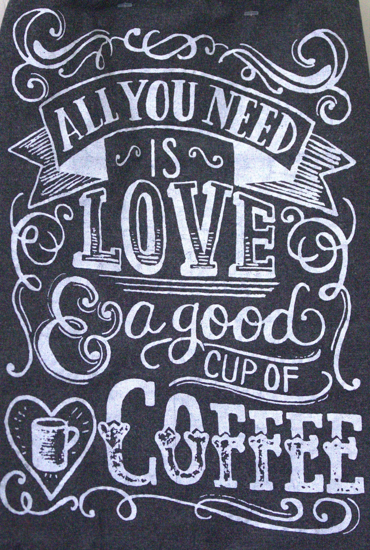 A close-up of a dark charcoal or black fabric printed with a white decorative typographic design that reads, 'ALL YOU NEED IS LOVE a good CUP OF COFFEE' with ornate flourishes and a coffee mug icon.
