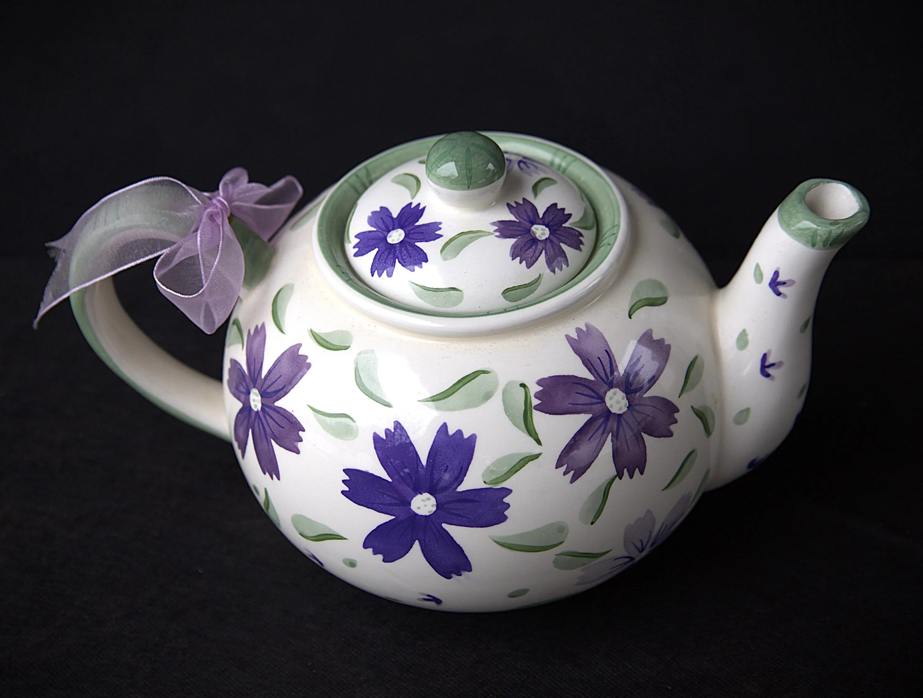 A white ceramic teapot decorated with purple flowers and green leaves, featuring a matching lid, a purple ribbon bow on the handle, and a black background.