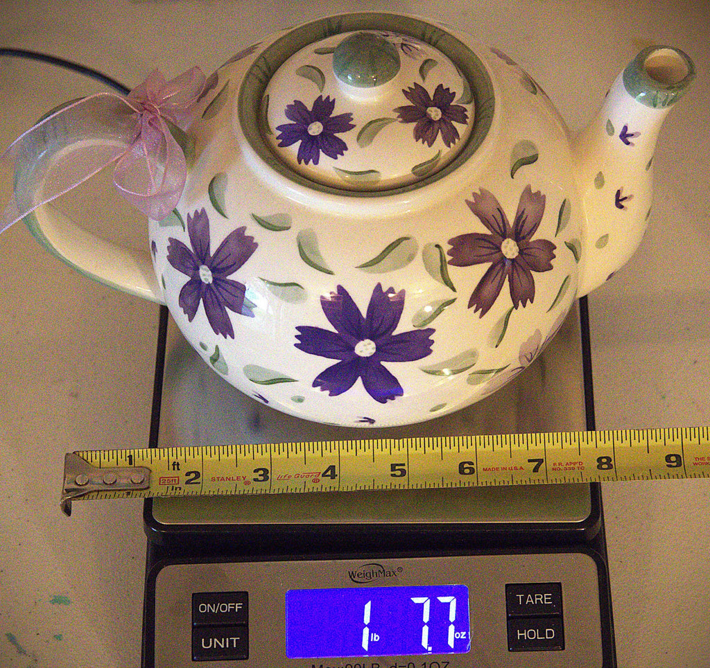 A white ceramic teapot decorated with purple flowers and green leaves, shown on a digital scale with a yellow measuring tape ruler underneath to indicate size. The teapot has a pink ribbon tied to the handle.