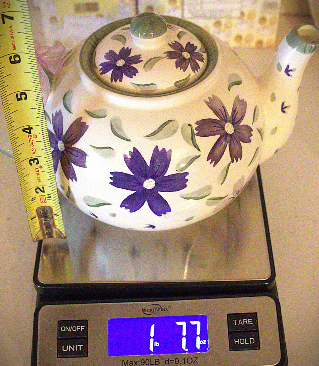 A white ceramic teapot decorated with purple flowers and green leaves, shown on a digital scale with a yellow measuring tape alongside it.