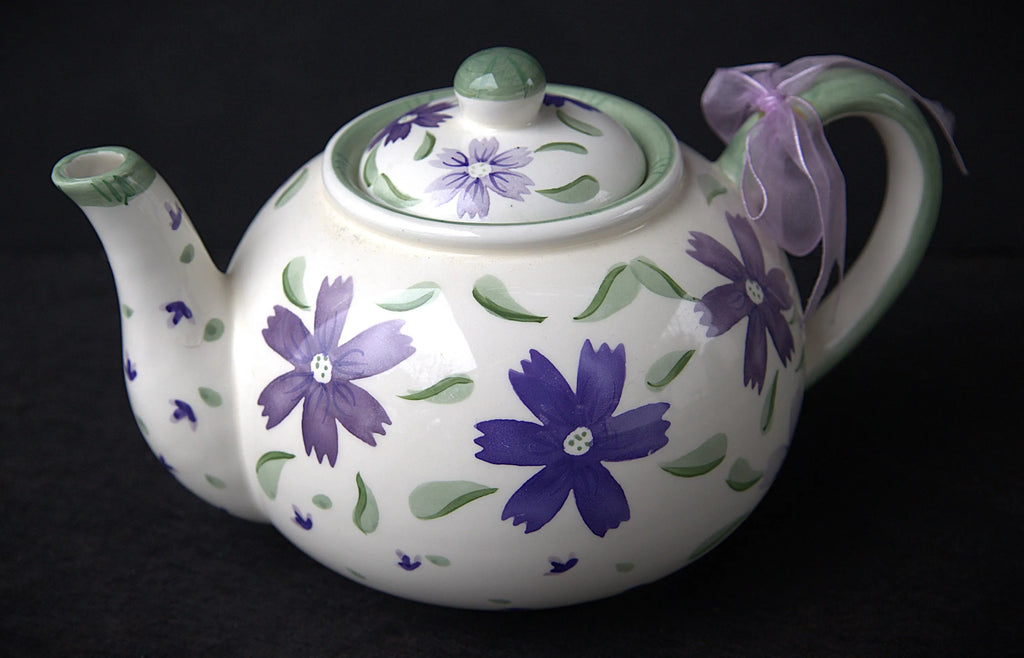 A white ceramic teapot decorated with purple flowers and green leaves, featuring a matching lid, green handle trim, and a light purple ribbon bow on the handle, set against a dark background.