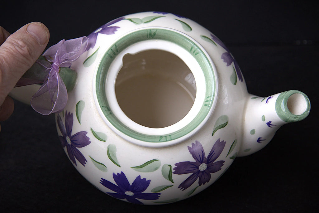 A hand holds a white ceramic teapot decorated with purple flowers and green leaves, featuring a green rim around the open top and a purple ribbon tied to the handle, set against a black background.