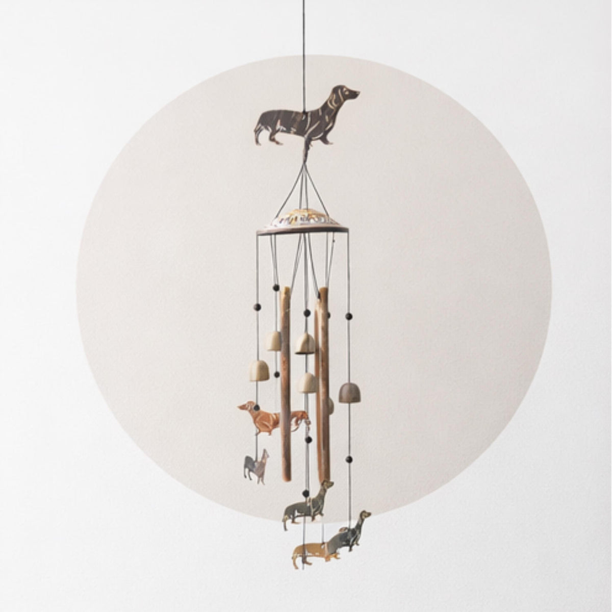 A hanging wind chime featuring a black silhouette of a dachshund at the top and several small animal figures below, with wooden tubes, small bells, and hanging weights, shown against a light gray background with a large pale circle behind it.