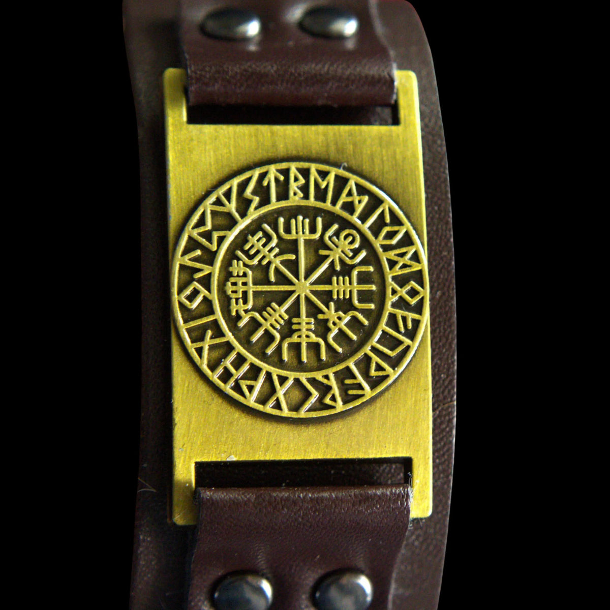 A close-up of a dark brown leather bracelet featuring a large rectangular gold-tone plate with a circular Viking ravenclaw symbol in the center, set against a solid black background.