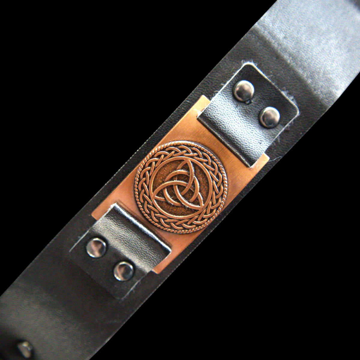 A close-up of a black leather belt featuring a central copper-toned metal plate with a Celtic knotwork triquetra design, set against a solid black background.