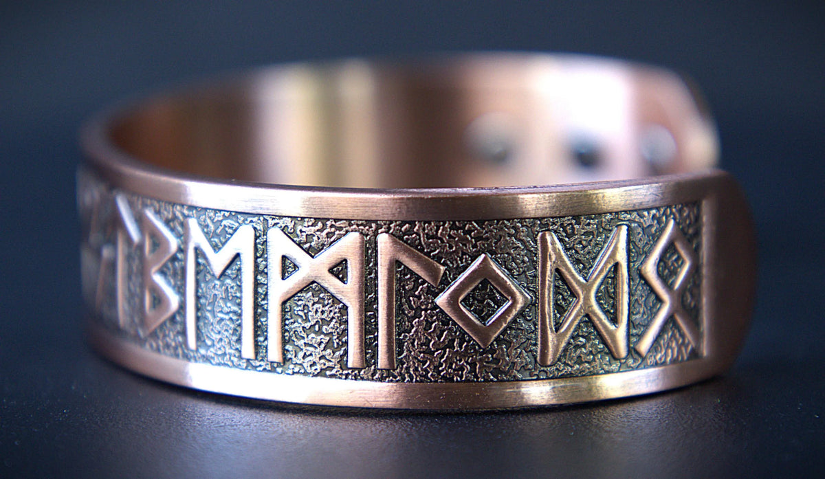 A close-up of a copper-colored metal cuff bracelet featuring engraved runic symbols on a textured band, set against a dark background.