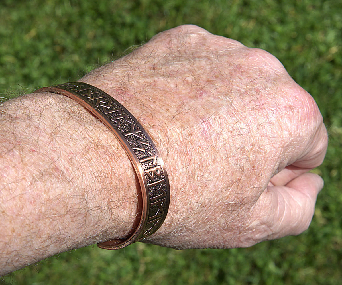 A close-up of a person's wrist wearing two copper-toned engraved bracelets against a blurred green grass background.