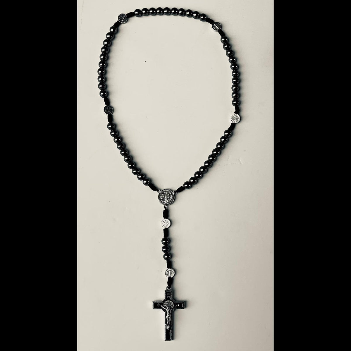 A black beaded rosary necklace with silver-toned metal details, including a crucifix pendant, displayed against a light beige background.