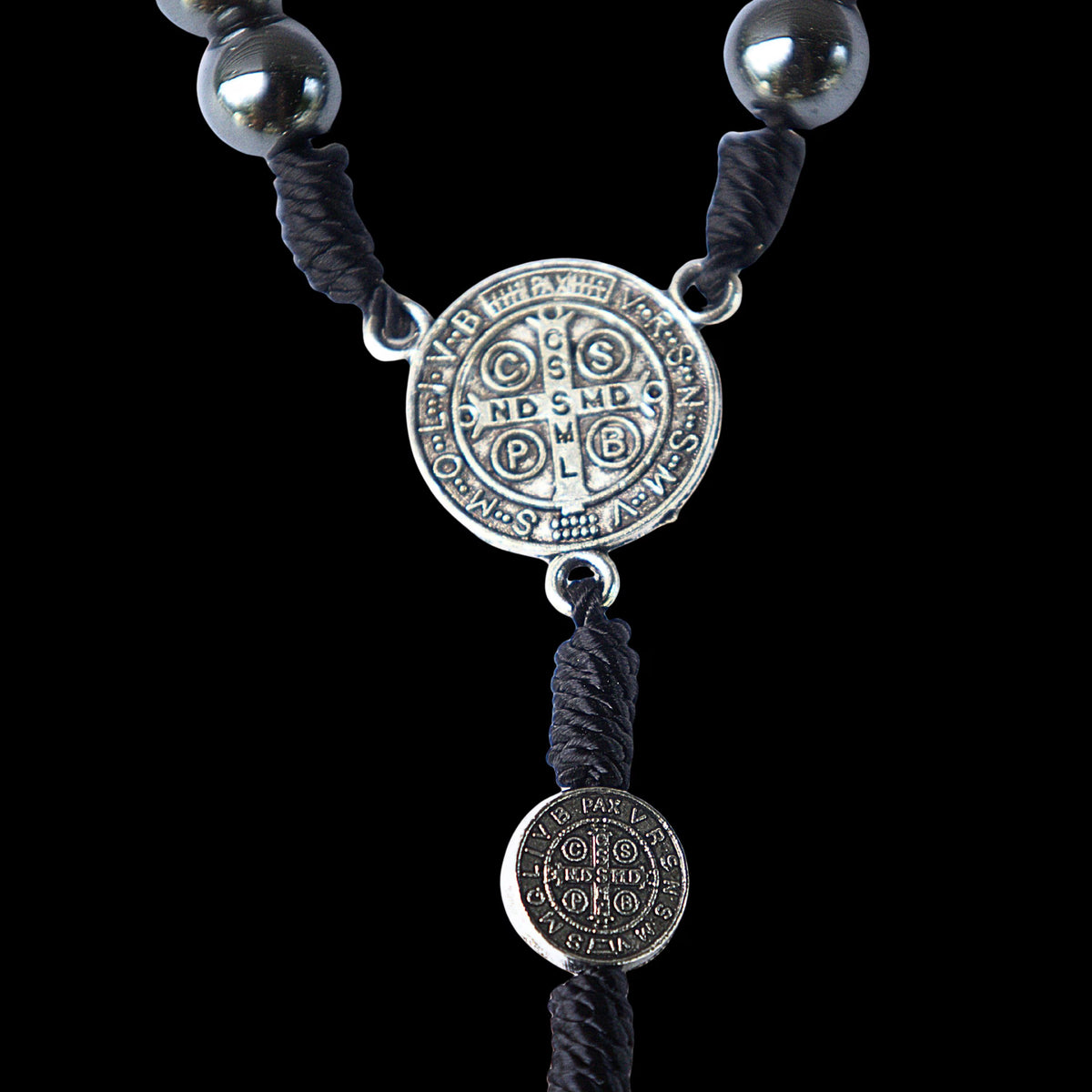 A close-up of a dark gray braided cord necklace with silver-toned metal beads and two round engraved medallion charms on a solid black background.
