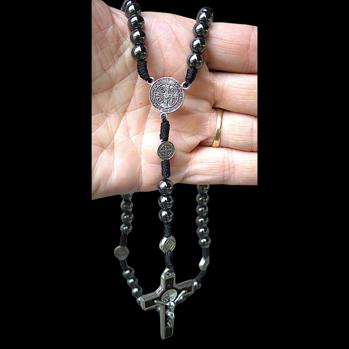 A black cord rosary necklace with dark metallic beads, silver-toned medallion pendants, and a crucifix, shown draped over a person's hand against a black background.