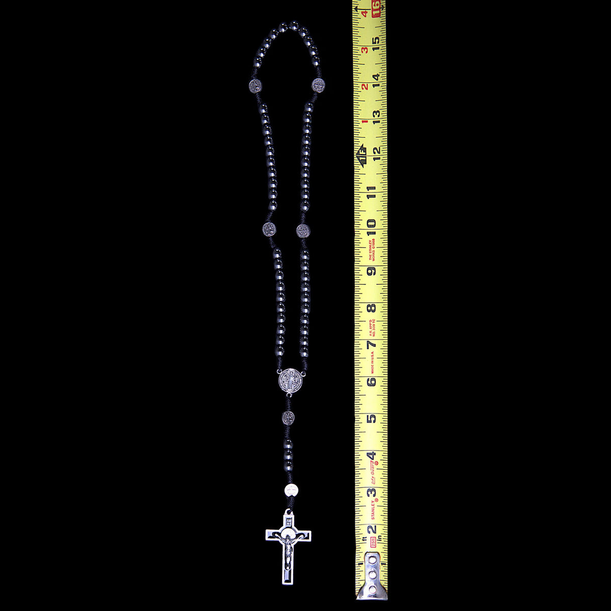 A black beaded rosary with silver-toned metal links, a silver crucifix pendant, and a yellow measuring tape standing vertically next to it against a solid black background.