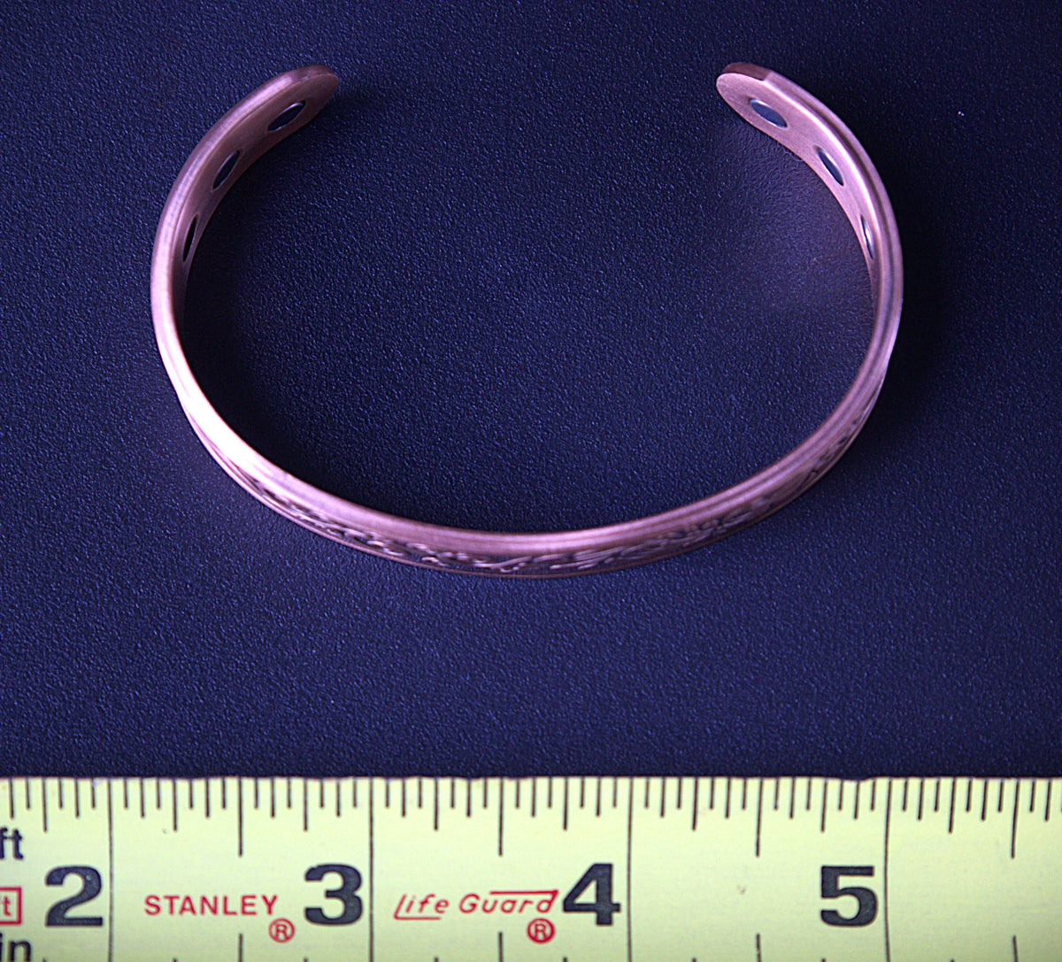 A pink metallic cuff bracelet with a textured pattern, displayed on a dark blue textured background with a yellow measuring tape ruler at the bottom for scale.