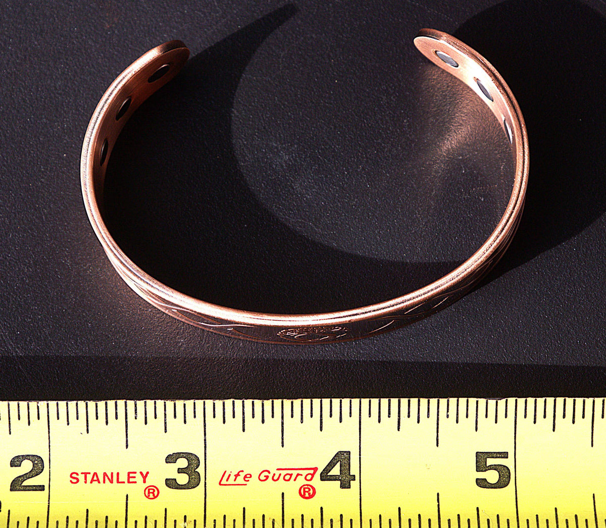 A copper-toned metal cuff bracelet with pre-drilled holes, shown next to a yellow Stanley Life Guard tape measure on a dark background.